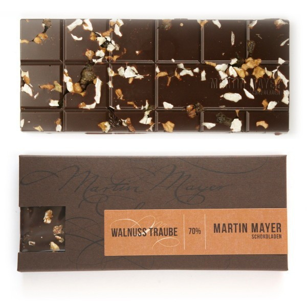 Verpackung und ausgepackte Tafel der Walnuss-Traube-Schokolade von Martin Mayer. Die Verpackung ist dunkelbraun mit rotbraunem Etikett und hat auf der kurzen Seite ein Sichtfenster, durch das man die Schokolade sehen kann. 