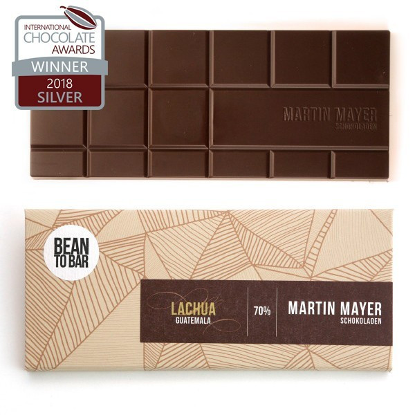 Verpackung und ausgepackte Tafel der Lachua-Schokolade von Martin Mayer. Die Verpackung ist beige mit dunkelbraunem Etikett und hat ein Muster aus hellbraunen alternierend schraffierten geometrischen Formen.