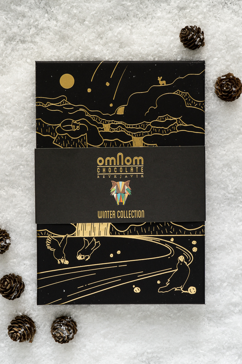 Verpackung der Omnom Winterkollektion - schwarze Schachtel mit goldenem Motiv von Winterlandschaft - auf Kunstschnee mit kleinen Tannenzapfen